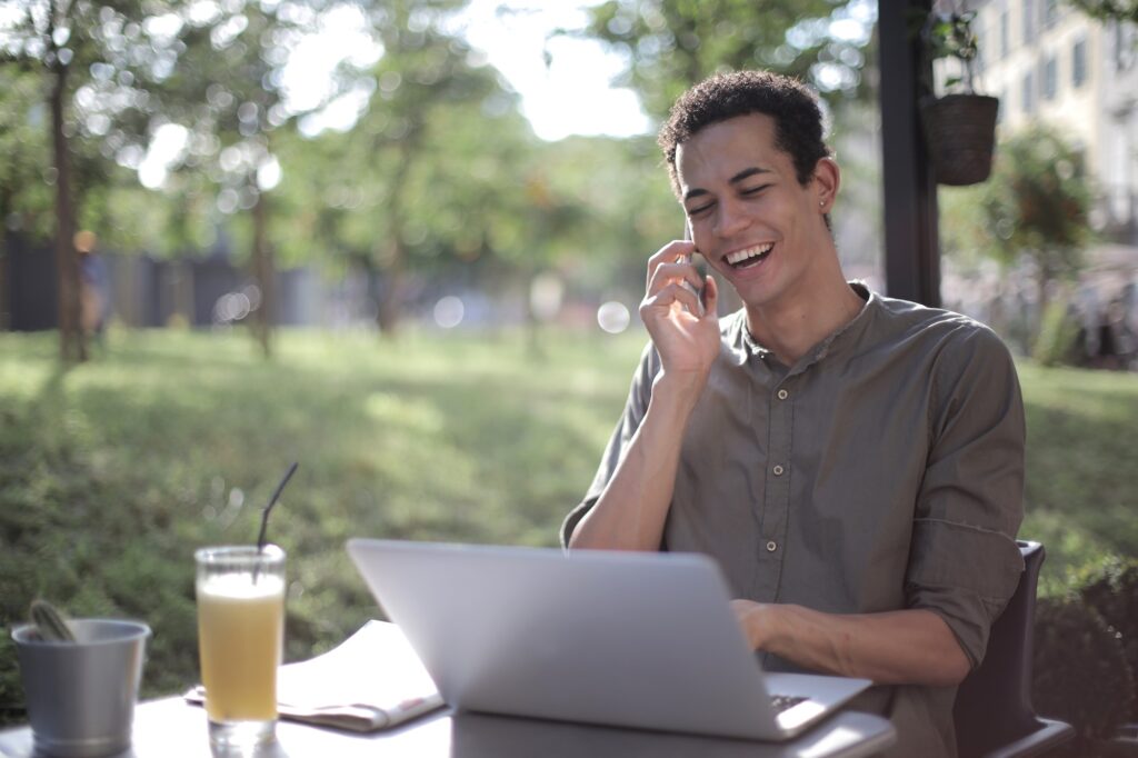 man talking on phone laughing in front of lap top with trees in the background
